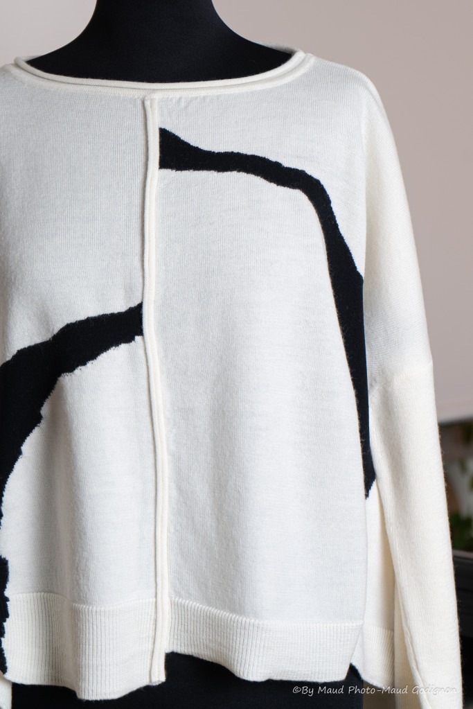 Pull ample et court écru avec motifs noirs. Composition bi-matière 100 % laine / 100 % nylon. Taille M. Photos des détails bas asymétrique et coutures apparentes.